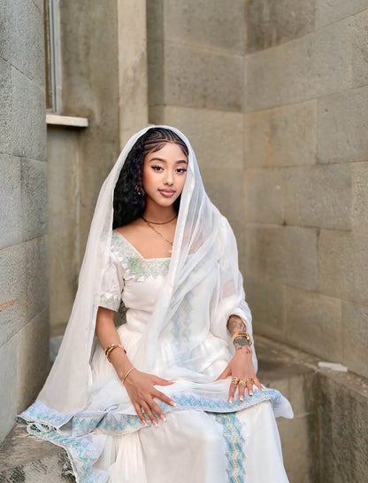 Woman in a white traditional Ethiopian cultural dress  with a veil sitting against a stone wall.
