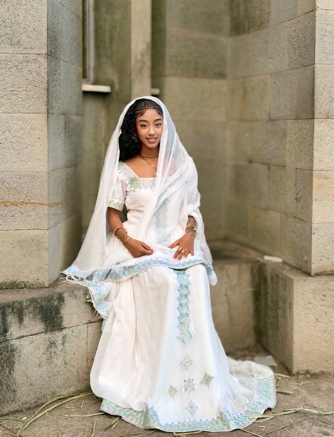 Woman in a white traditional Ethiopian cultural dress sitting against a stone wall.