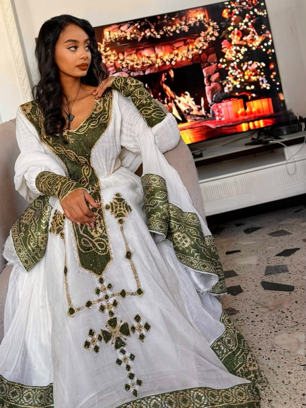 Woman in a white and green embroidered Ethiopian dress sitting in front of a TV displaying a festive scene.