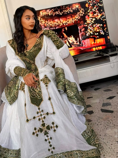 Woman in a white and green embroidered Ethiopian dress sitting in front of a TV displaying a festive scene.