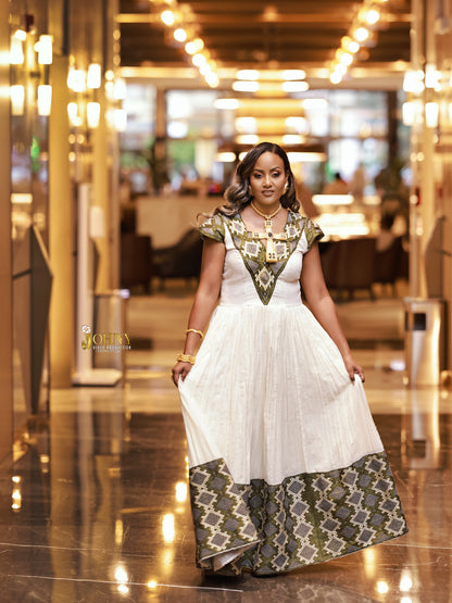A woman modeling a white African-style Ethiopia Clothes with intricate green and gold patterned fabric details on the bodice, sleeves, and hem, standing in a brightly lit indoor hallway.