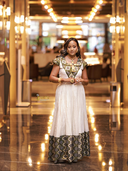 A woman stands in a grand, warmly lit hotel lobby wearing a traditional Ethiopian long Ethiopia Clothes with a white body, gold and green patterned sleeves and hem, and gold jewelry.