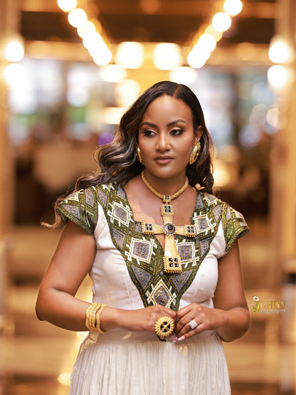 A woman wearing a white Ethiopia Clothes with an ornate green and gold geometric embroidered yoke, styled with gold jewelry in a warmly lit indoor setting.