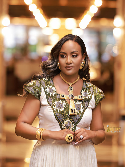 A woman wearing a white Ethiopia Clothes with an ornate green and gold geometric embroidered yoke, styled with gold jewelry in a warmly lit indoor setting.