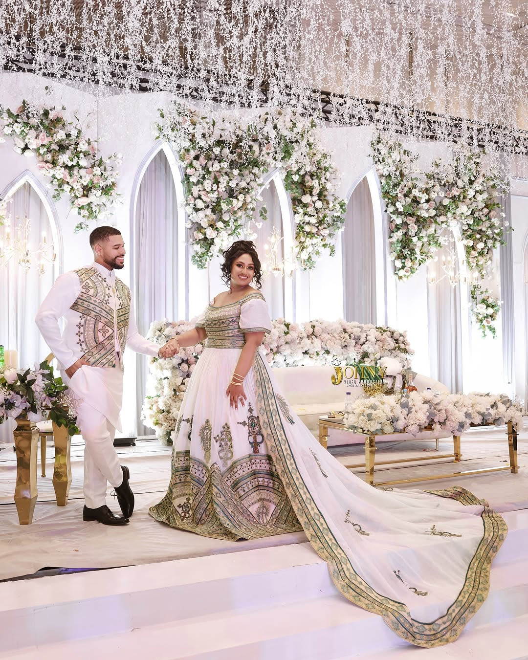 Man and woman in formal Habesha Outfit couple Set  standing in a decorated indoor setting with floral arrangements.