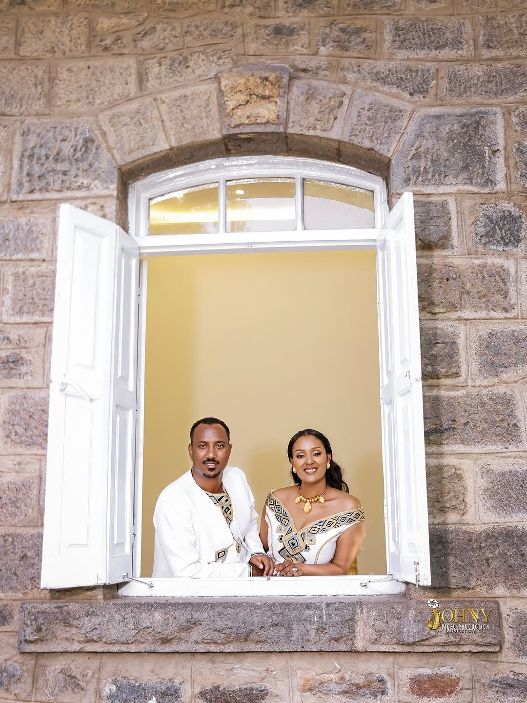 Couple in Family Habesha Set  standing behind an open window with a stone wall background