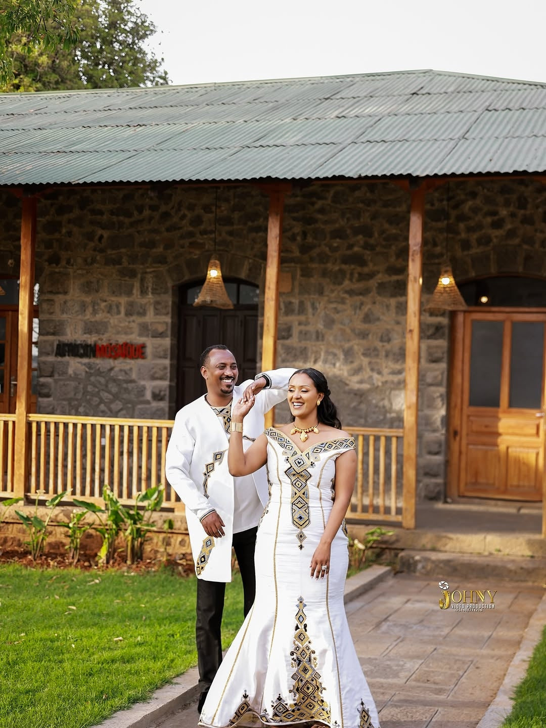 Couple in Family Habesha Set attire standing in front of a stone building with a wooden porch.