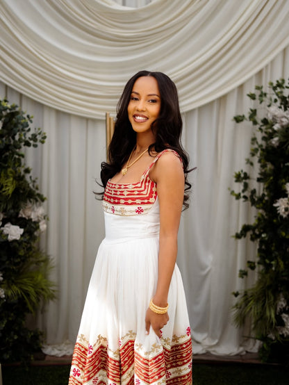 Woman in a white and red patterned Ethiopian cultural wedding dress  standing in front of a decorative backdrop with plants.