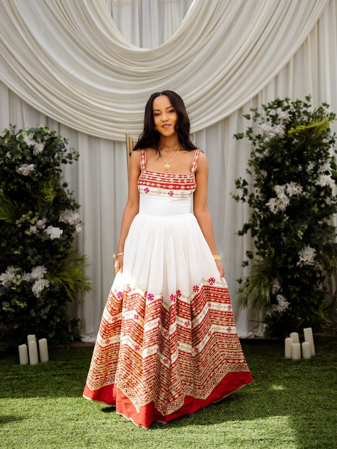Woman in a white and red patterned Ethiopian cultural wedding dress standing in front of decorative white curtains and greenery.