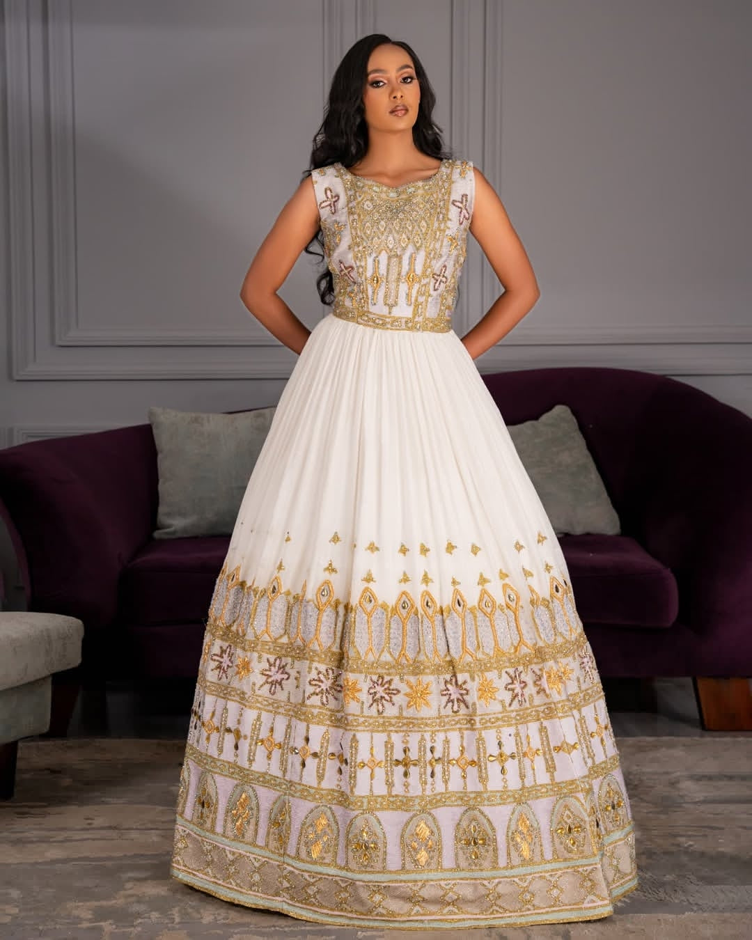 Woman wearing a white and gold Ethiopian wedding dress in a room with a purple couch.