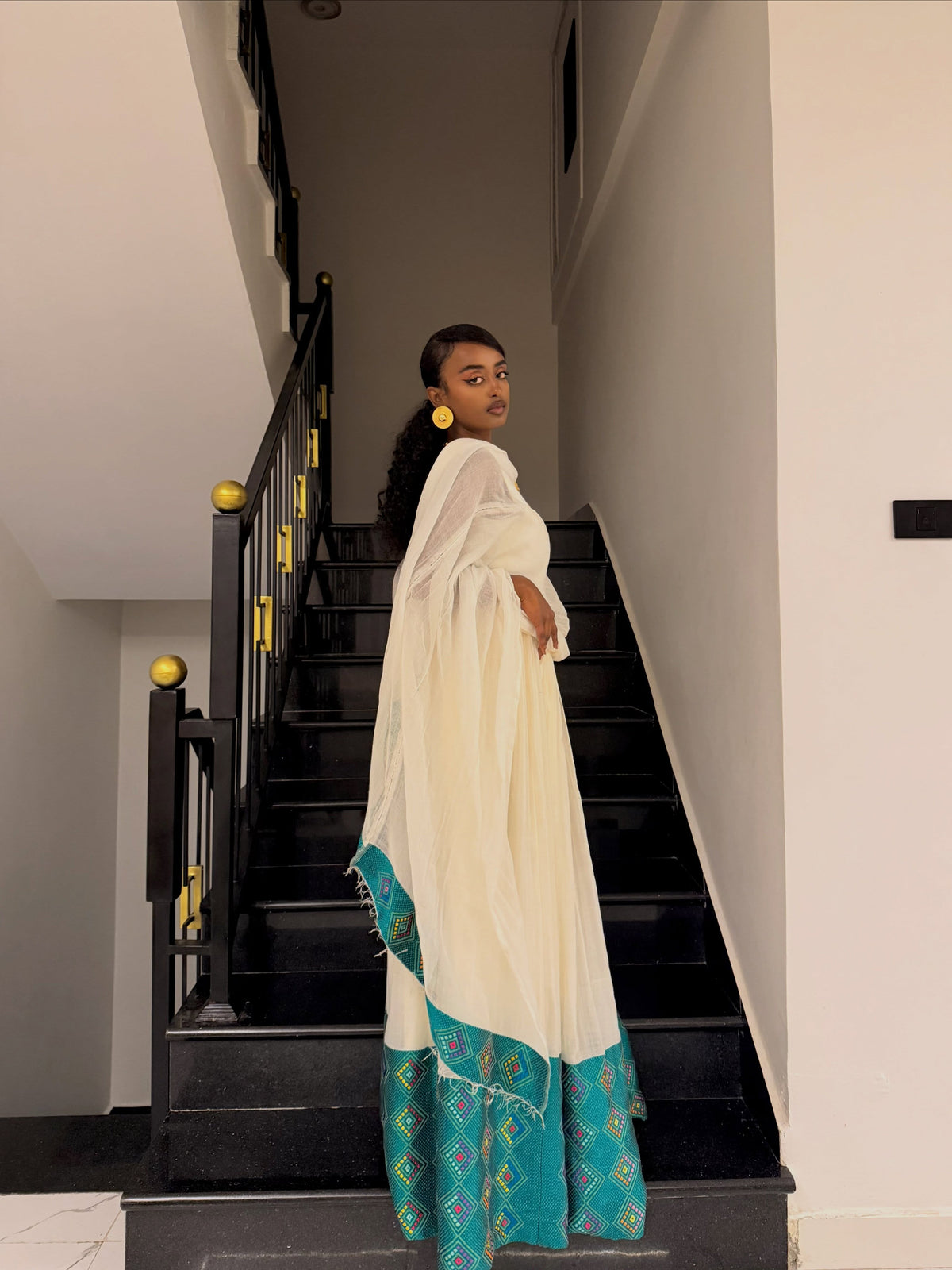 A woman wearing a traditional white and teal embroidered Ethiopian Traditional Dress stands on a black staircase indoors, looking back over her shoulder. The dress has a solid white upper portion and a teal lower portion with intricate geometric embroidery.