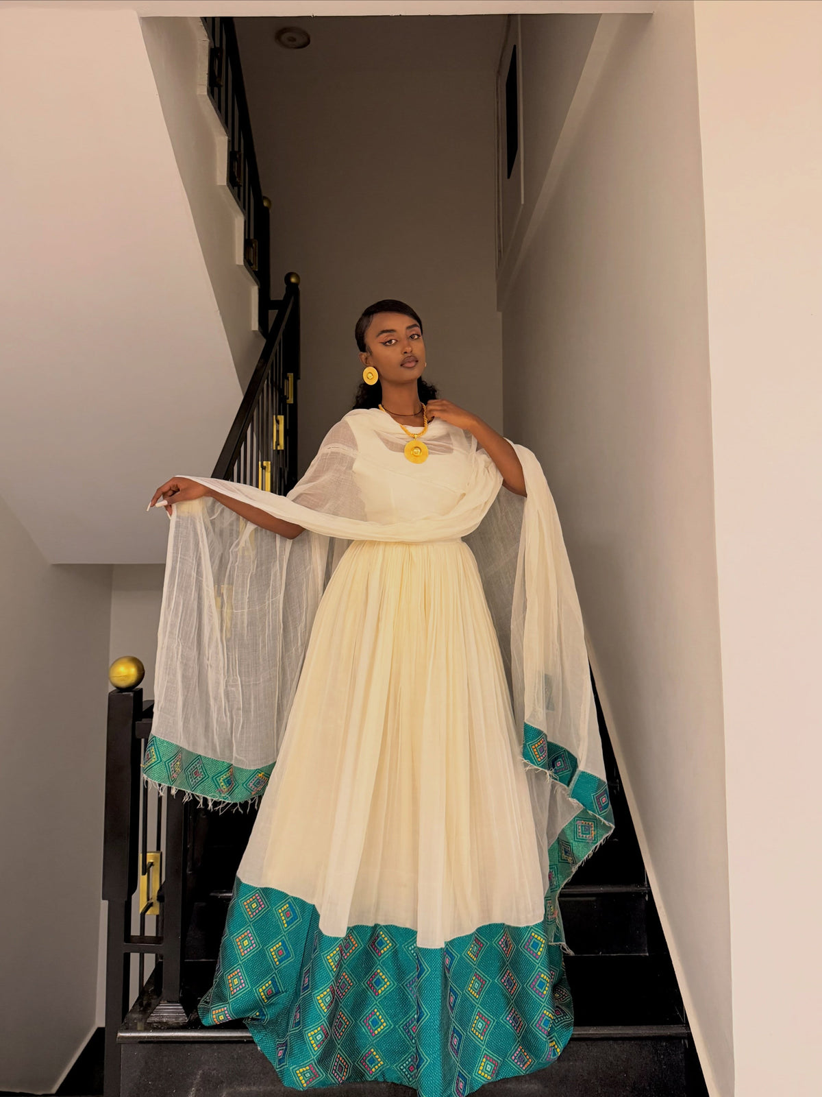 A woman stands on an indoor staircase wearing a traditional Ethiopian Traditional Dress consisting of a cream-colored dress with a teal patterned hem and matching sheer dupatta, accessorized with gold jewelry.