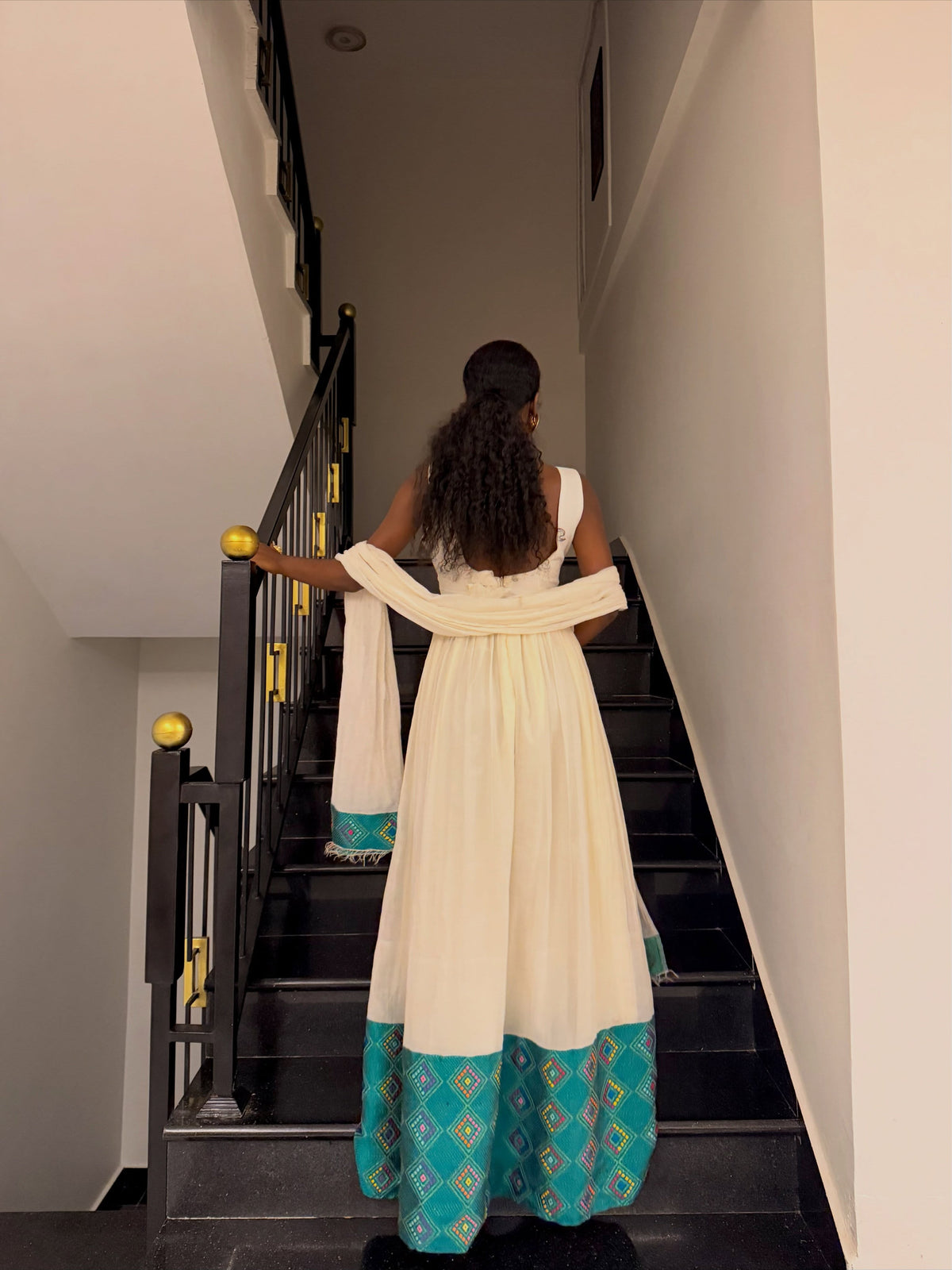 A woman viewed from behind wearing a cream-colored traditional Ethiopian Traditional Dress with a teal and gold patterned hem, walking up a black staircase indoors.