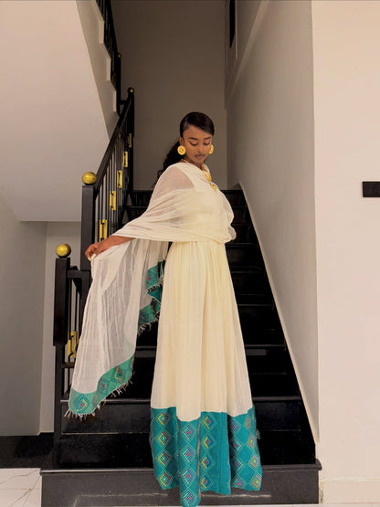A woman wearing a traditional white and teal Ethiopian Traditional Dress with a sheer white dupatta stands on a black staircase indoors.