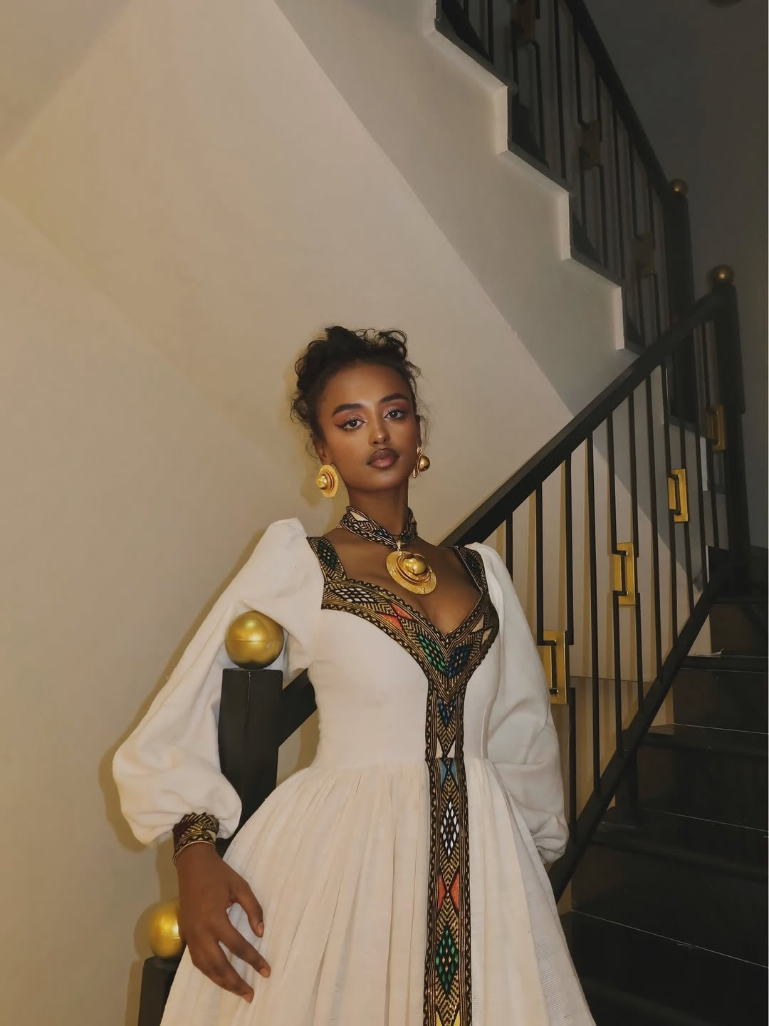 Woman in a white Habesha Libs  with gold accessories standing in a staircase.