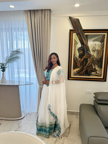 Woman in a white and blue traditional Ethiopian Traditional Dress  standing in a modern living room with a painting on the wall.