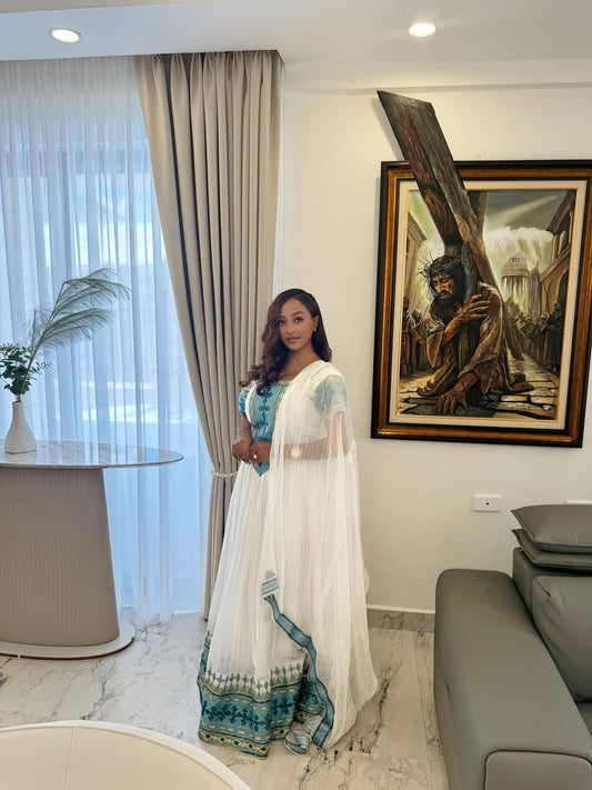 Woman in a white and blue traditional Ethiopian Traditional Dress  standing in a modern living room with a painting on the wall.