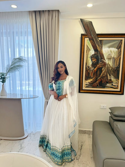Woman in a white and blue traditional Ethiopian Traditional Dress  standing in a modern living room with a painting on the wall.