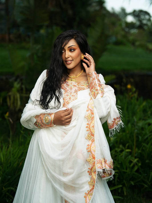 A woman wearing a white traditional Ethiopian shop near me with orange and gold embroidered details, standing outdoors in a lush green garden setting.