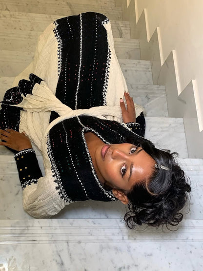 A woman lying on white marble stairs wearing a black and cream Ethiopian Dress  with white embroidered stripes and black knit cuffs.
