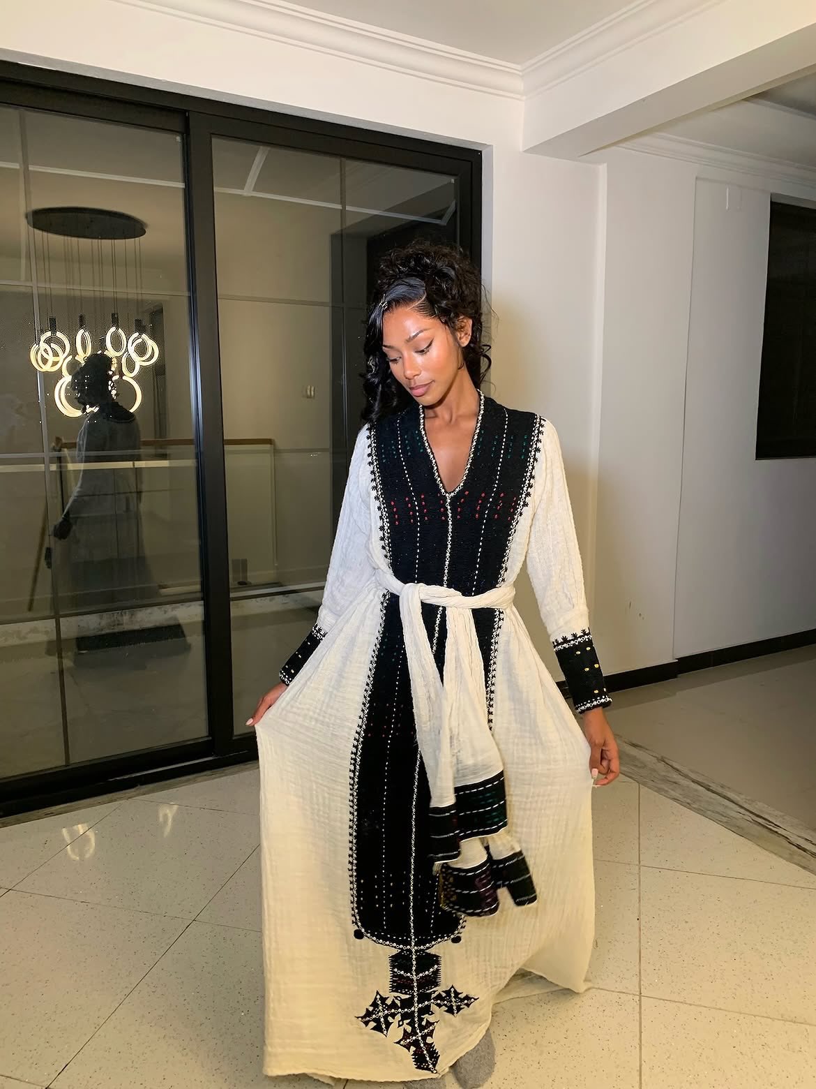 A woman wearing a long white Ethiopian Dress 
with black embroidered details and a matching white belt, standing in a modern indoor setting with glass doors.