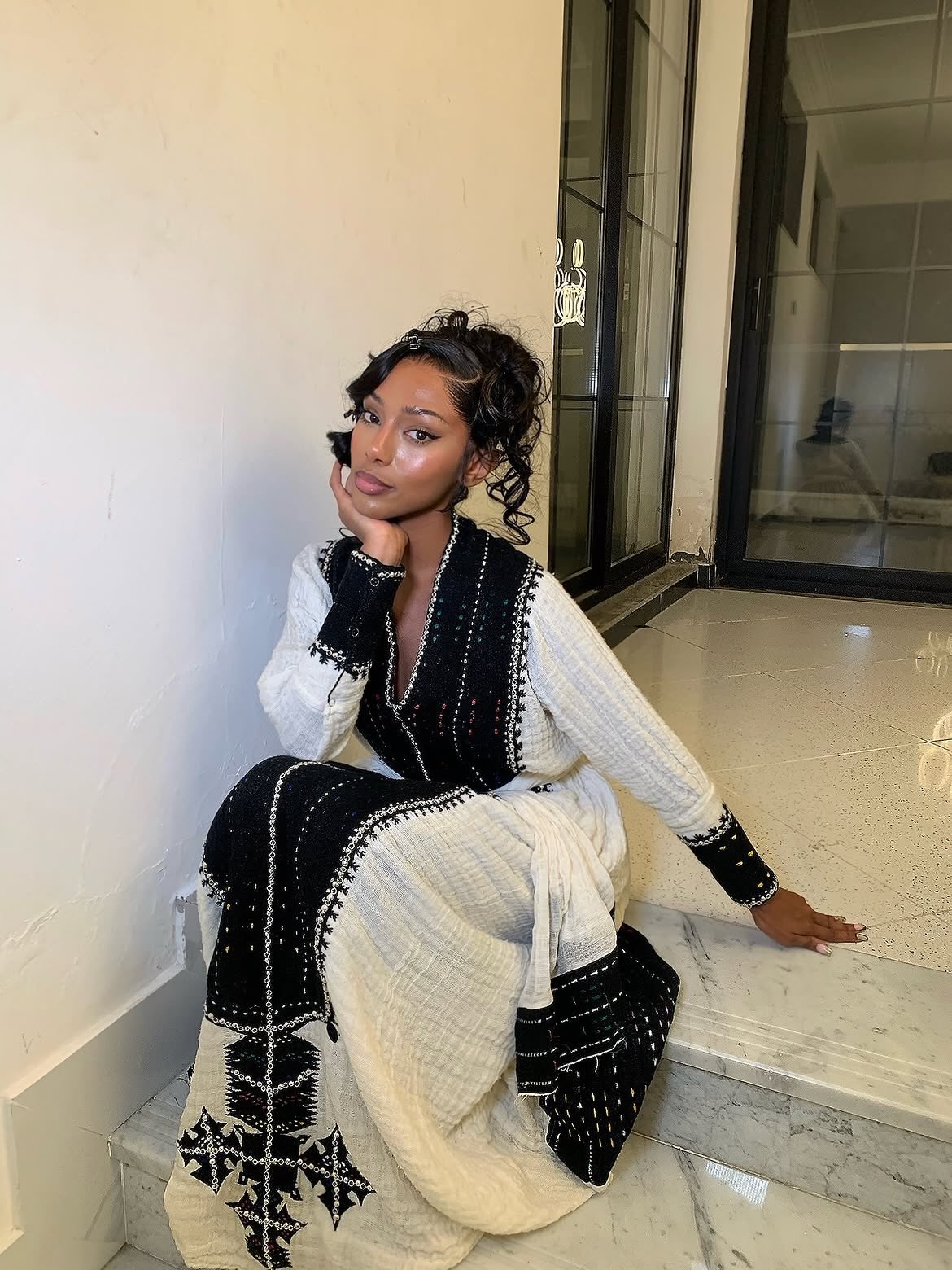 A woman sits on marble steps indoors wearing a long white Ethiopian Dress  with black embroidered patterns and black sleeve cuffs.