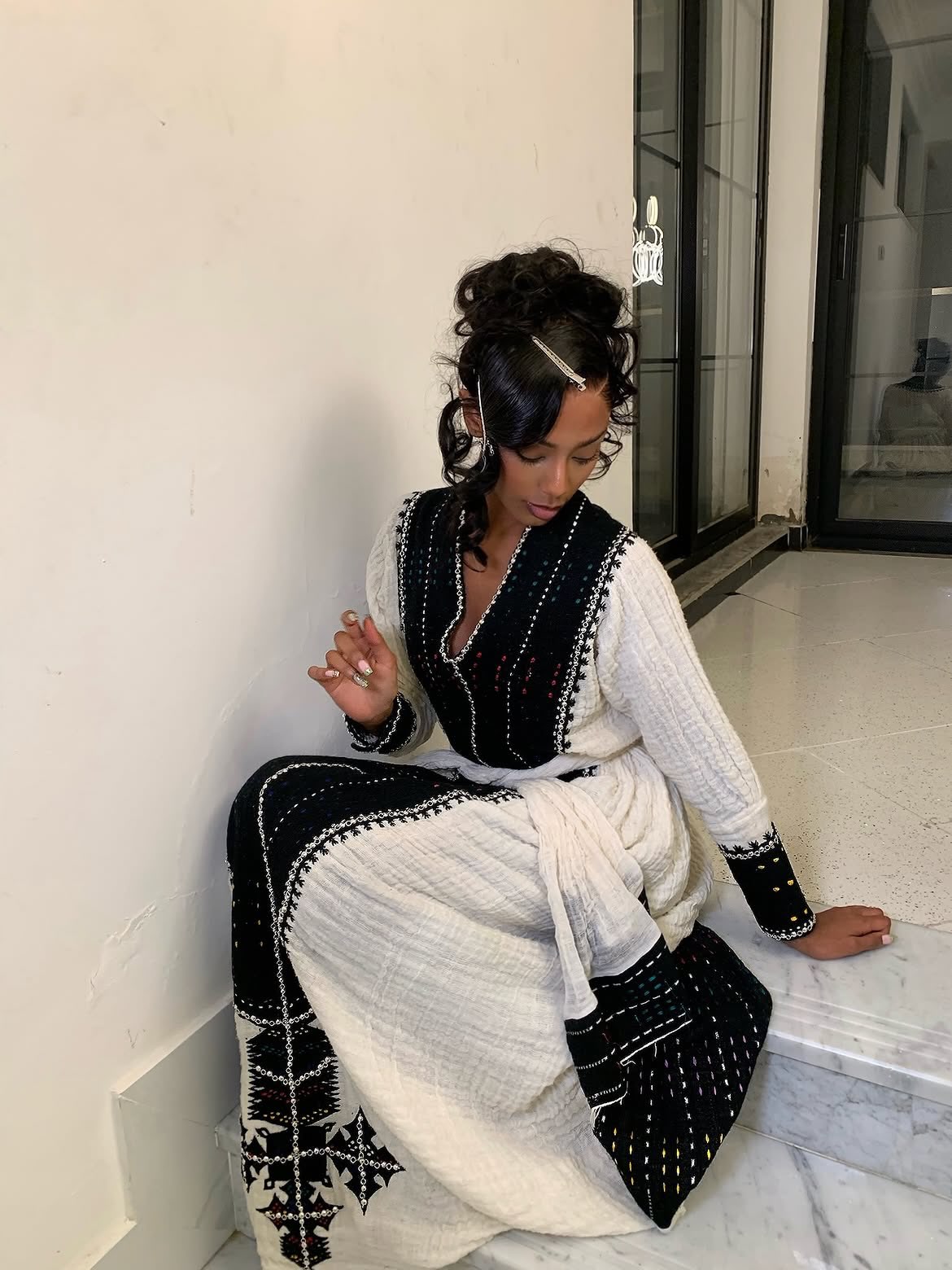 A woman sits on a marble ledge wearing a black and white embroidered traditional Ethiopian Dress  with a white textured outer layer and black embroidered panels.