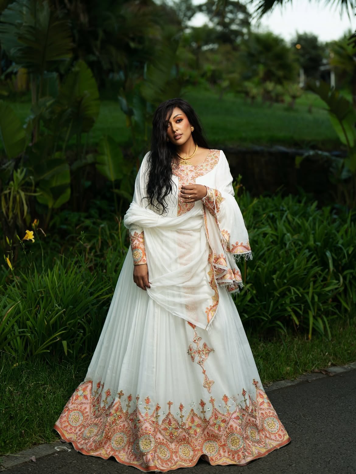 A woman wearing a white traditional Ethiopian shop near me with peach and gold embroidery, standing outdoors in a lush green garden setting.