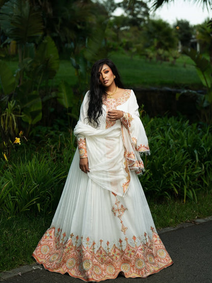 A woman wearing a white traditional Ethiopian shop near me with peach and gold embroidery, standing outdoors in a lush green garden setting.