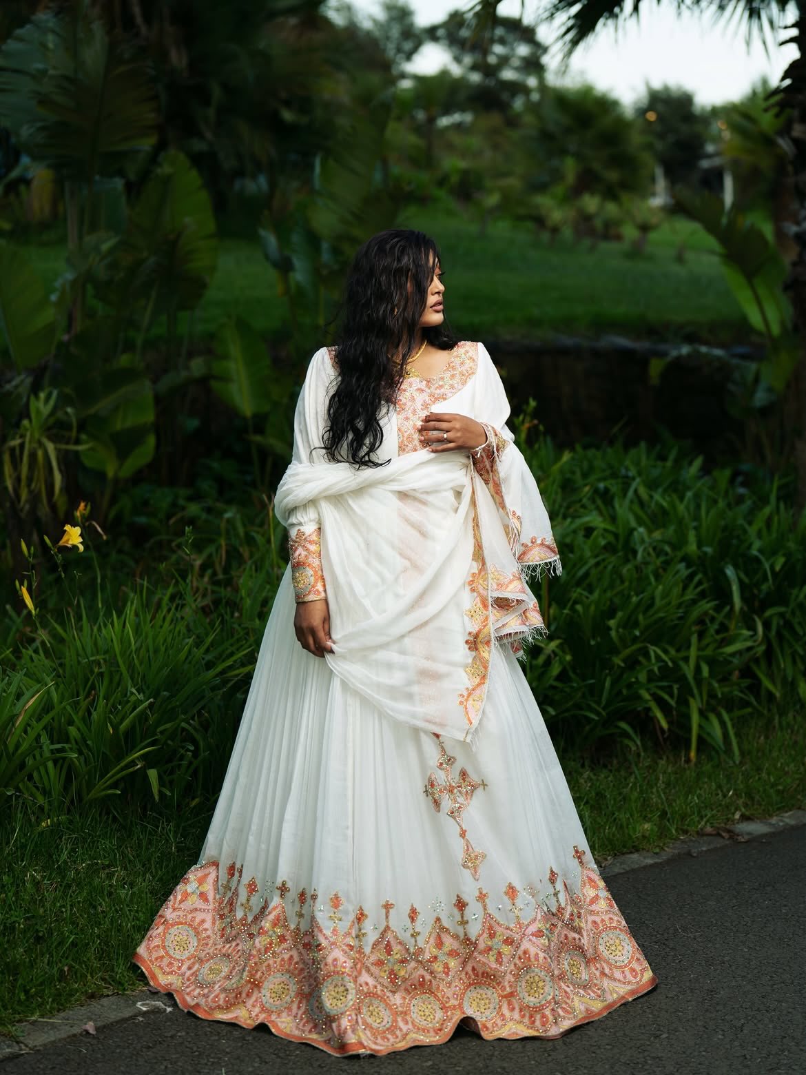 A woman wearing a white traditional Ethiopian shop near me with peach and gold floral embroidery, standing outdoors on a paved path surrounded by lush green tropical foliage.