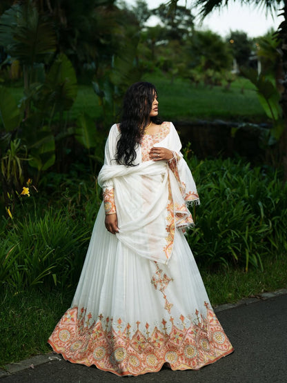 A woman wearing a white traditional Ethiopian shop near me with peach and gold floral embroidery, standing outdoors on a paved path surrounded by lush green tropical foliage.