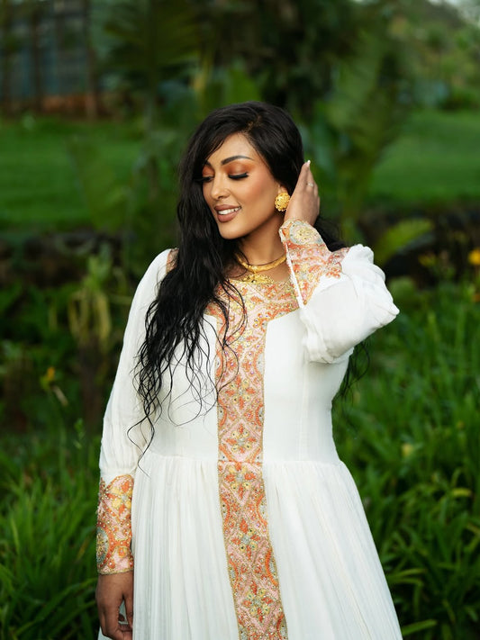 A woman wearing a white long-sleeve Ethiopian shop near me with a peach and green paisley patterned front panel and matching patterned cuffs, standing outdoors in a lush green garden.