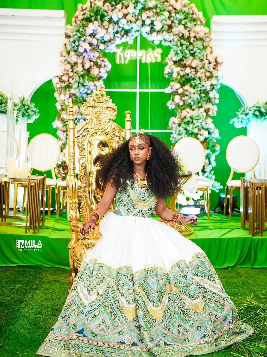 Woman in a green and white traditional Green Habesha Wedding Dress sitting on a decorated throne with floral arches.