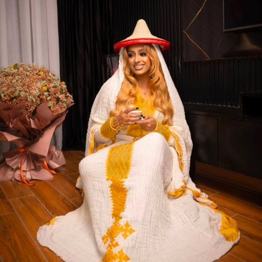 Person in a Yellow Eritrean Dress with a hat, sitting indoors with a bouquet of flowers on the side.
