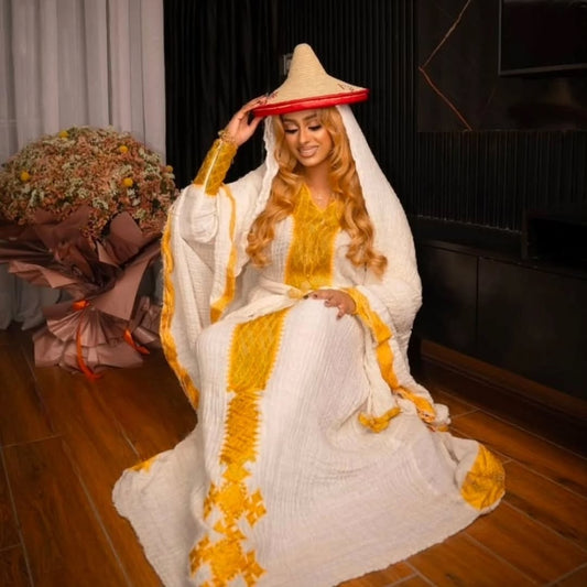 Woman in a white and yellow traditional Yellow Eritrean Dress with a large hat indoors.