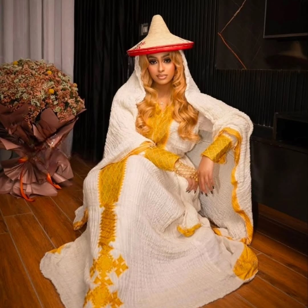 Person in a Yellow Eritrean Dress with a cone-shaped hat sitting on a wooden floor.