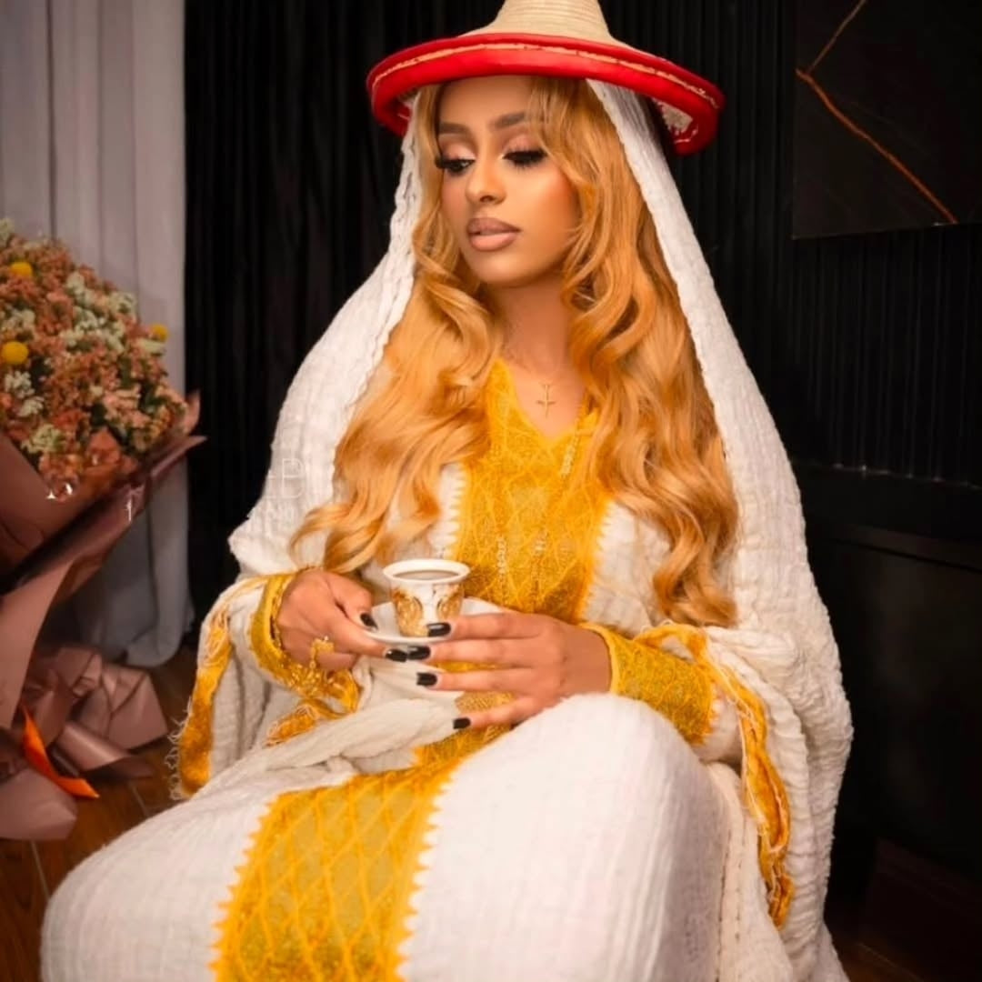 Woman in a yellow and white Yellow Eritrean Dress with a red hat, holding a cup.