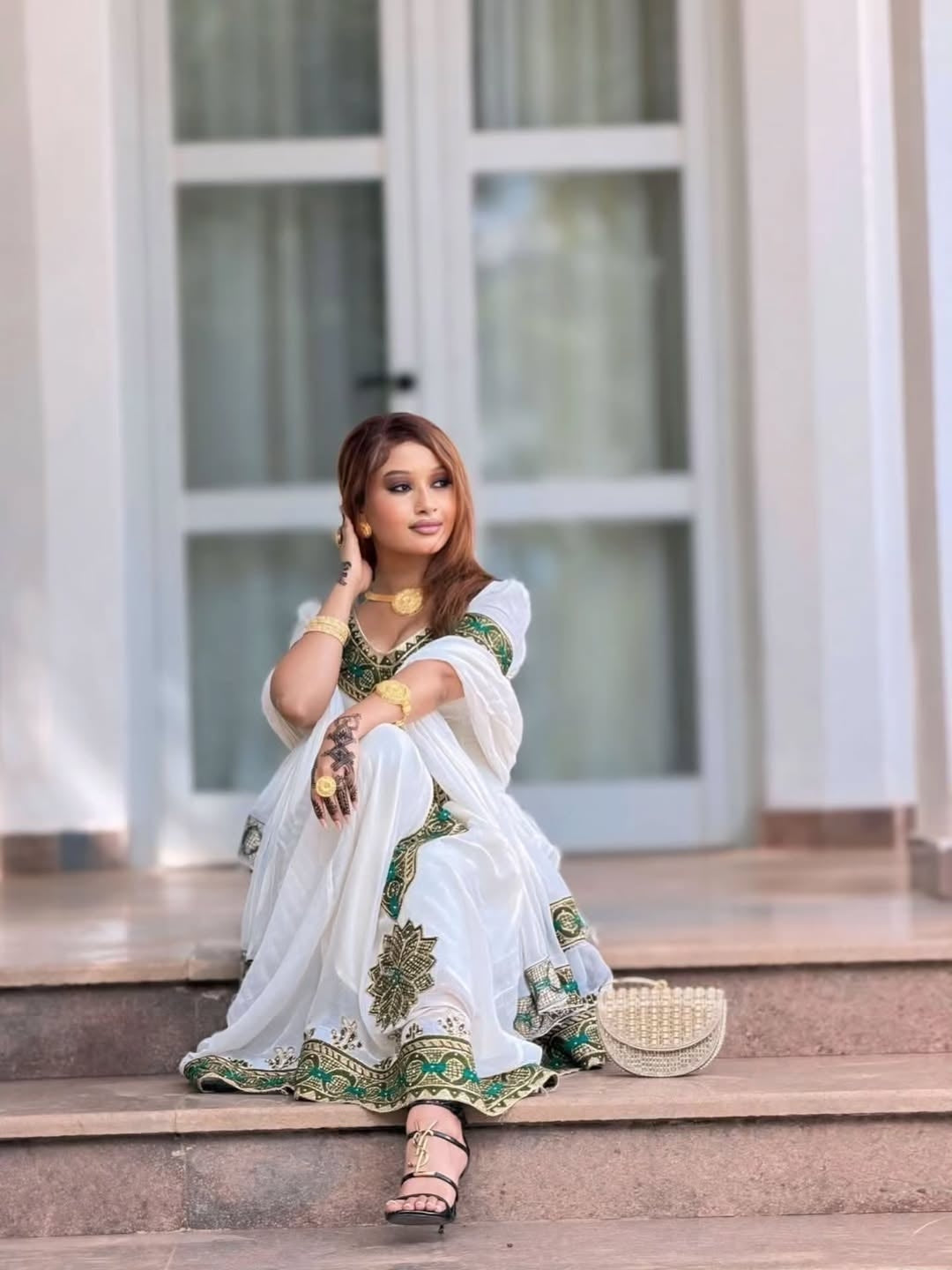 Woman in a white saree with green patterns sitting on steps.
