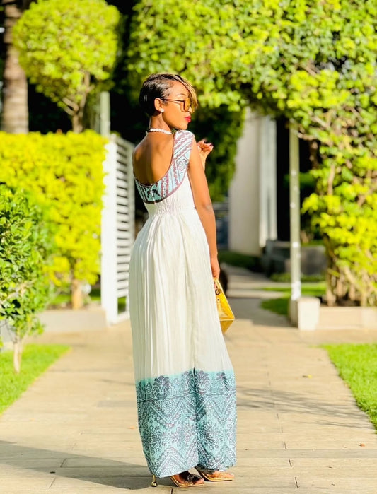Woman in a long, patterned dress standing on a sidewalk with greenery in the background