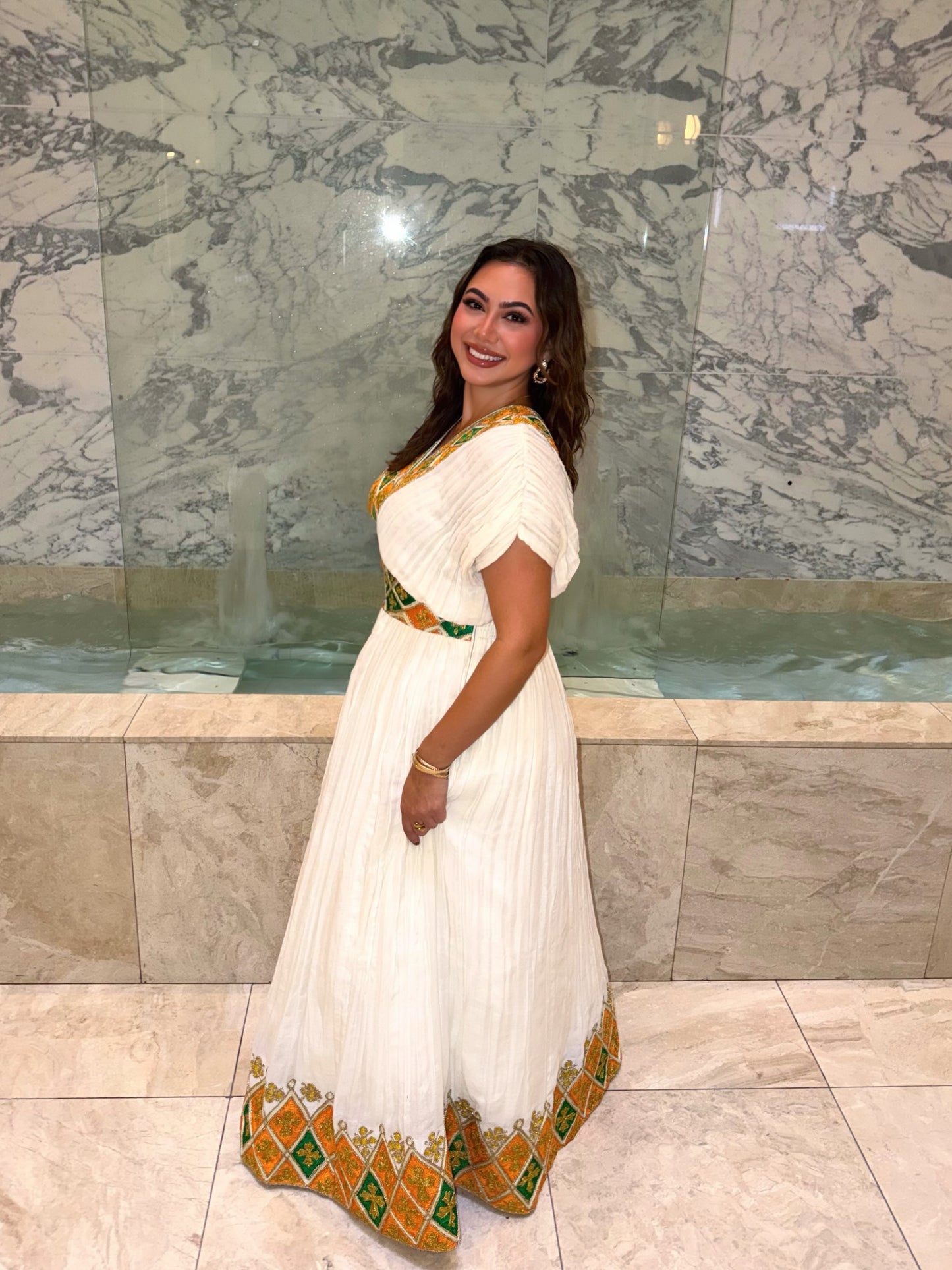 A woman modeling a white pleated  Habesha Dress with gold and green geometric border details, standing in front of a marble wall and pool.