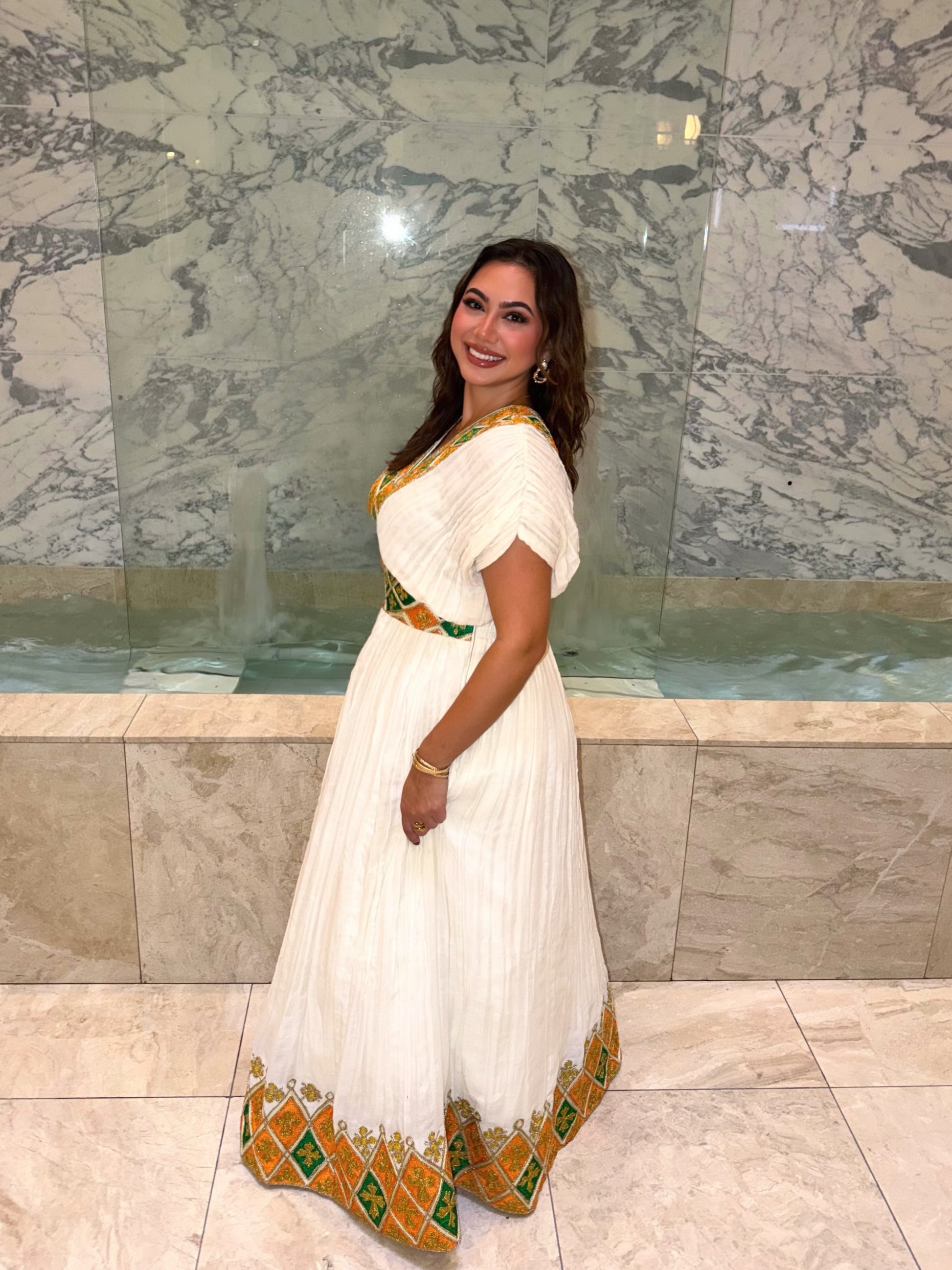 A woman modeling a white pleated  Habesha Dress with gold and green geometric border details, standing in front of a marble wall and pool.