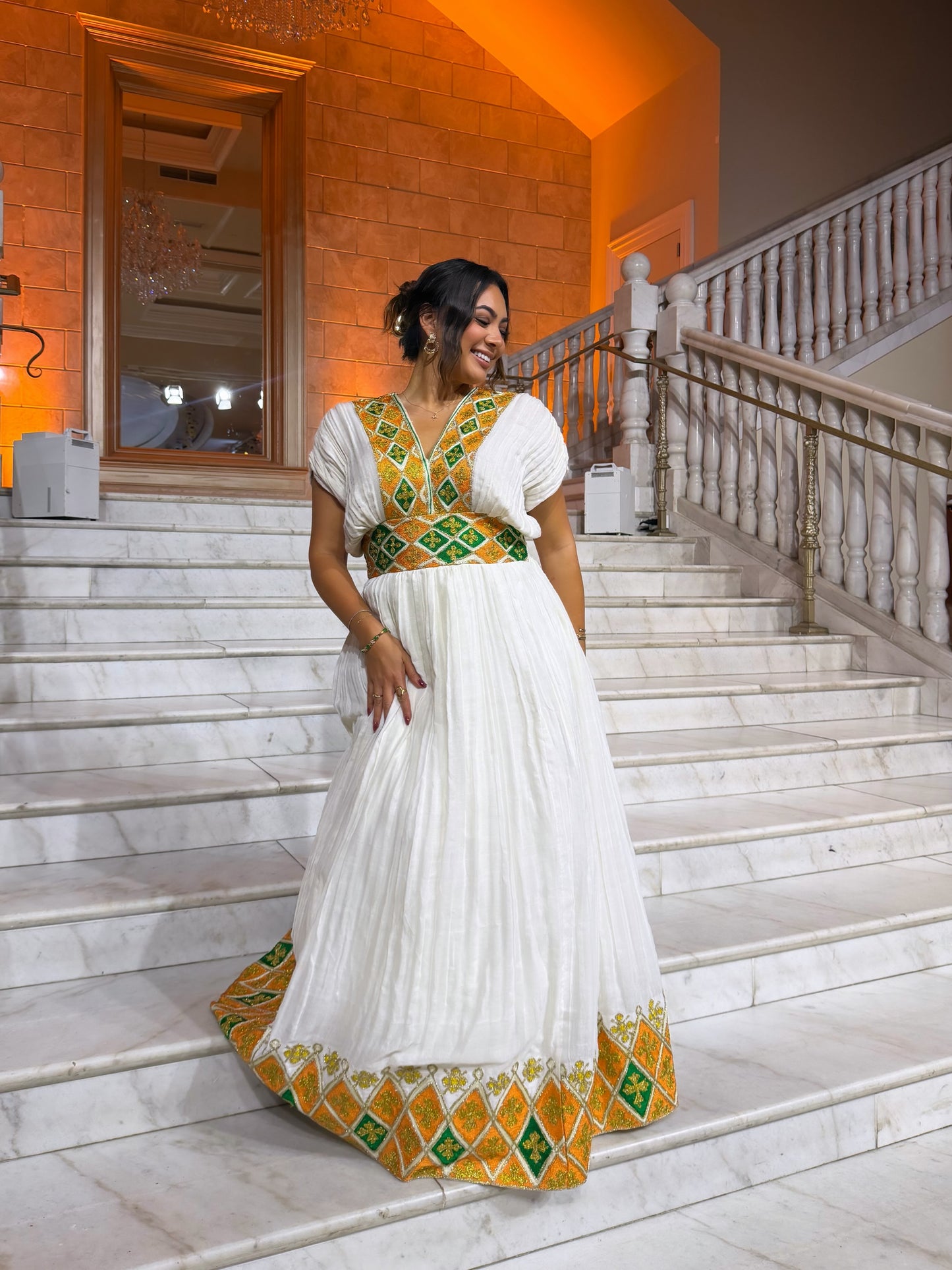 A woman wearing a white traditional  Habesha Dress with green and gold geometric embroidery, standing on a marble staircase in a grand indoor venue with warm orange lighting.