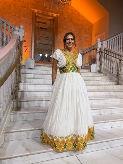 A woman wearing a traditional white  Habesha Dress with green and yellow geometric embroidery, standing on a grand marble staircase in a warmly lit building.