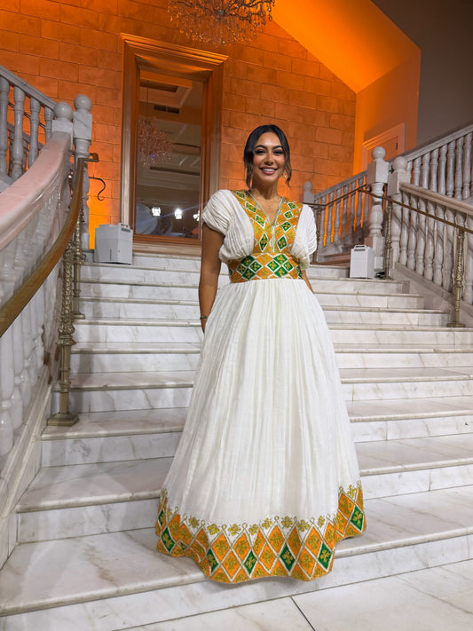 A woman wearing a traditional white  Habesha Dress with green and yellow geometric embroidery, standing on a grand marble staircase in a warmly lit building.