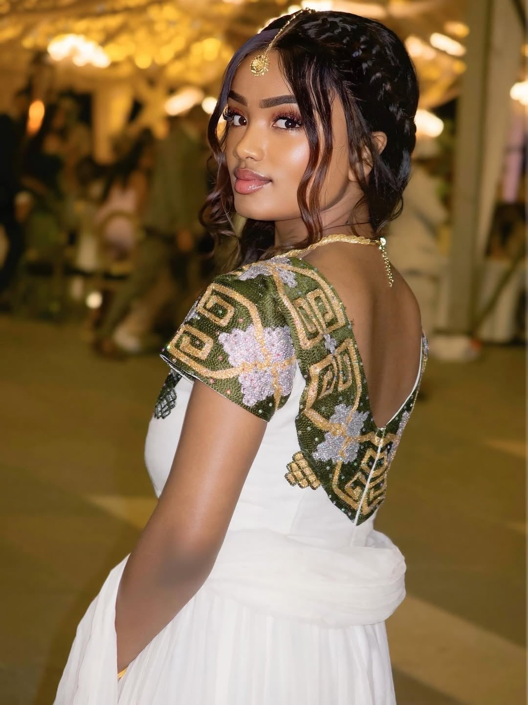 Woman wearing a traditional habesha dress with floral and geometric patterns in a blurred indoor setting