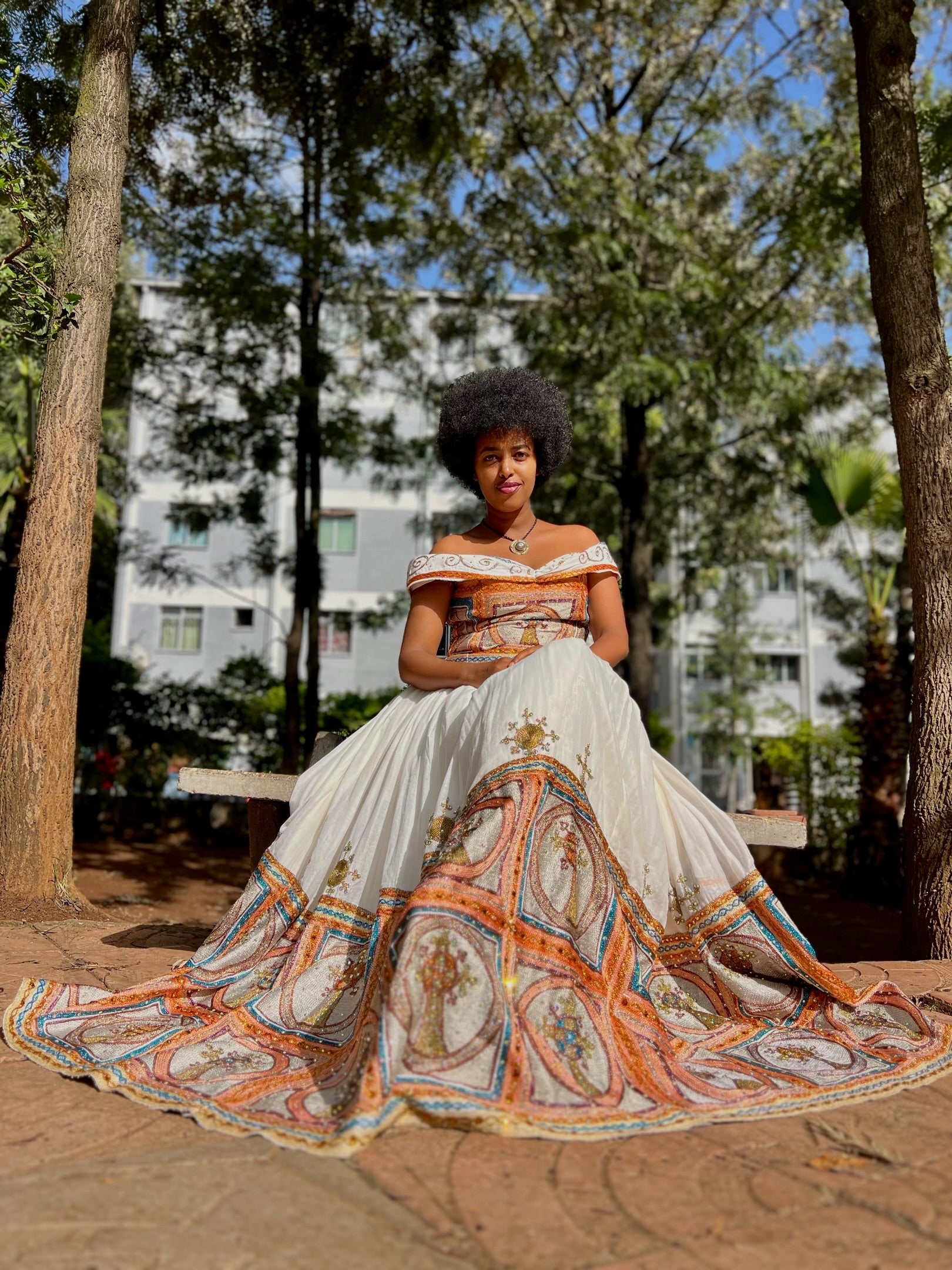 Woman in a white and orange patterned dress standing outdoors with trees and buildings in the background.