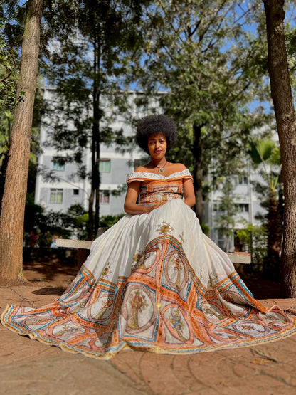 Woman in a white and orange patterned dress standing outdoors with trees and buildings in the background.