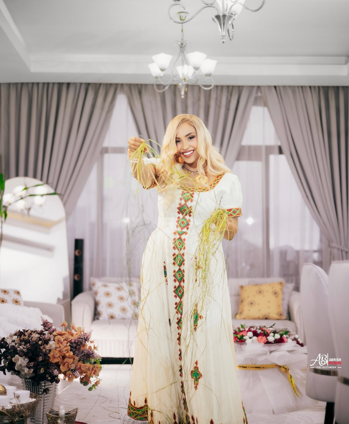 A woman with long blonde wavy hair wearing a whitehabesha kemis with colorful geometric embroidery in red, green, and gold, accessorized with gold necklaces and bracelets, posing indoors.