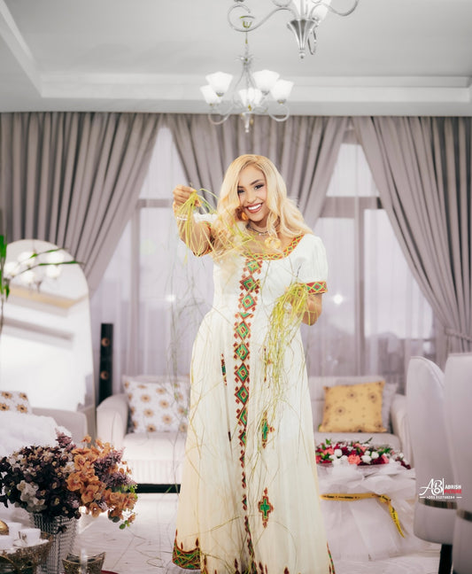 A woman with long blonde wavy hair wearing a whitehabesha kemis with colorful geometric embroidery in red, green, and gold, accessorized with gold necklaces and bracelets, posing indoors.