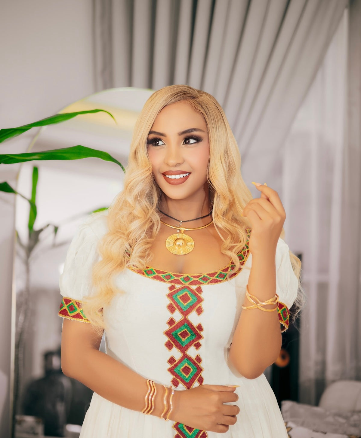 A woman with long blonde wavy hair wearing a white habesha kemis with colorful geometric embroidery in red, green, and gold, accessorized with gold necklaces and bracelets, posing indoors.